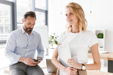 Paperwork ile ayakta işbirliği işçisi kadın ile ofiste bir masa üzerine yaslanmış iken cep telefonunuzu kullanarak kendine güvenen adam Yöneticisi