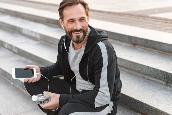 Su şişesi açık havada adımlarla üzerinde otururken cep telefonuyla kulaklık sporcu gülümseyen