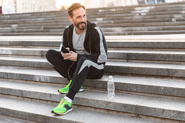 Su şişesi açık havada adımlarla üzerinde otururken cep telefonuyla kulaklık mutlu sporcu