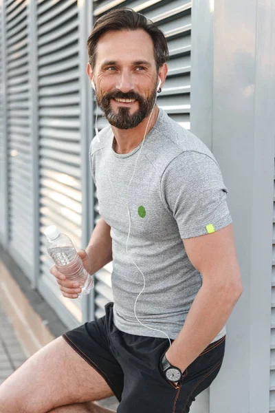 İmge-in yakışıklı mutlu güçlü olgun sporcu ayakta açık havada şişe su ile tutarak kulaklık ile bir kenara dinleme müzik arıyorum.