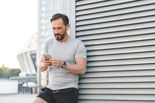 İmge-in yakışıklı güçlü olgun sporcu ayakta açık havada cep telefonuyla earphones ile bir kenara dinleme müzik arıyorum.