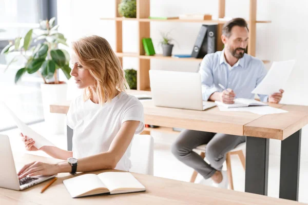 背景に男の同僚とオフィスで座っている間ドキュメントをラップトップ コンピューターに取り組んで集中して女性マネージャー — ストック写真