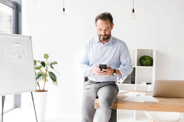 Hombre Negocios Sonriente Usando Teléfono Móvil Mientras Apoya Escritorio Oficina — Foto de Stock