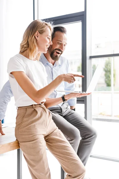 Memnun meslektaş ofis pencerenin önünde duran ve parmak işaret ederken dizüstü bilgisayar üzerinde çalışan birkaç