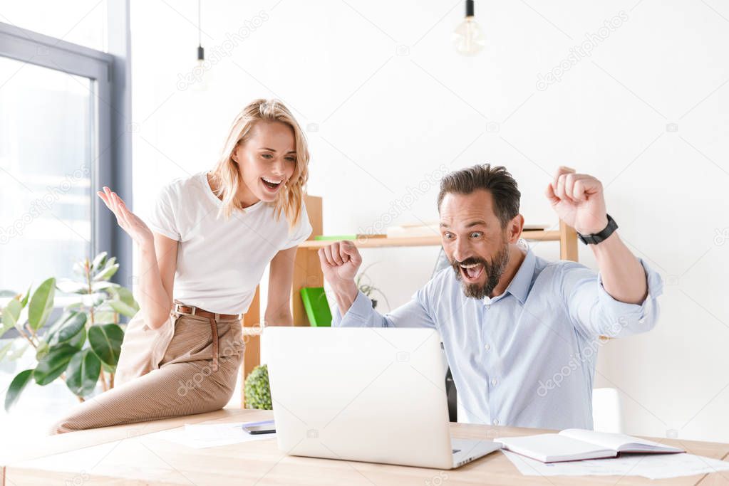 Couple of happy colleagues celebrating success while sitting at the office and looking at laptop computer