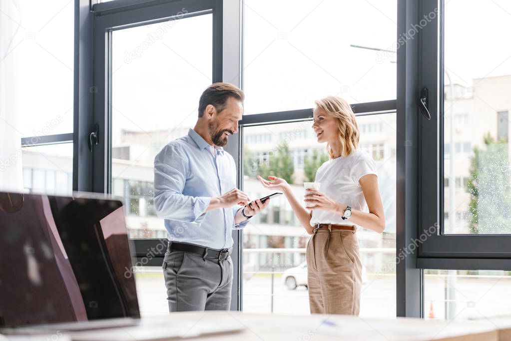 Couple of excited colleagues talking while standing at the office window with mobile phone and cup of coffee