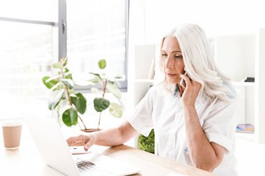Kapalı ofis çalışma dizüstü bilgisayar kullanan cep telefonuyla konuşurken oturan Olgun kadın fotoğrafı.