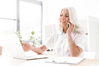 Neşeli pozitif Olgun kadın kapalı ofis çalışma dizüstü bilgisayar kullanan cep telefonuyla konuşurken oturan görüntüsünü.