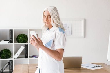 Kapalı ofis çalışma içme içinde olgun mutlu kadın cep telefonuyla kahve görüntüsünü.