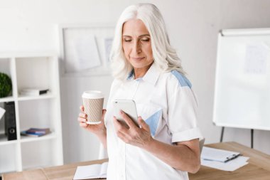 Kapalı ofis çalışma içme içinde olgun mutlu kadın cep telefonuyla kahve görüntüsünü.