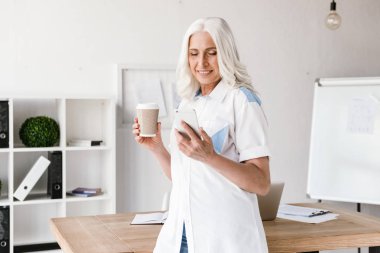 Kapalı ofis çalışma içme içinde olgun mutlu kadın cep telefonuyla kahve görüntüsünü.