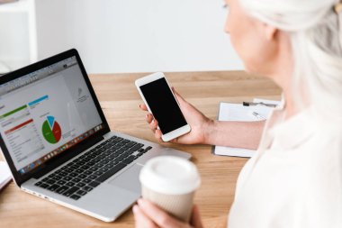 Dizüstü bilgisayarda çalışarak ve Office masada oturan ve cep telefonu tutan kahve içme emin olgun iş kadını, yakın çekim
