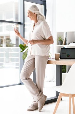 Gülümseyen olgun iş kadını ofiste masasında duran cep telefonu kullanarak