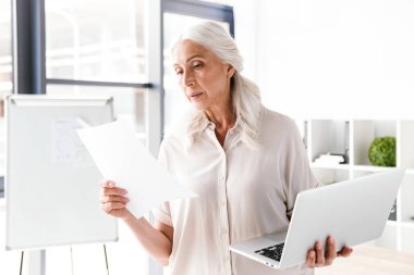 Emin olgun iş kadını ofiste ayakta ve dizüstü bilgisayar tutan belgeleri analiz etme