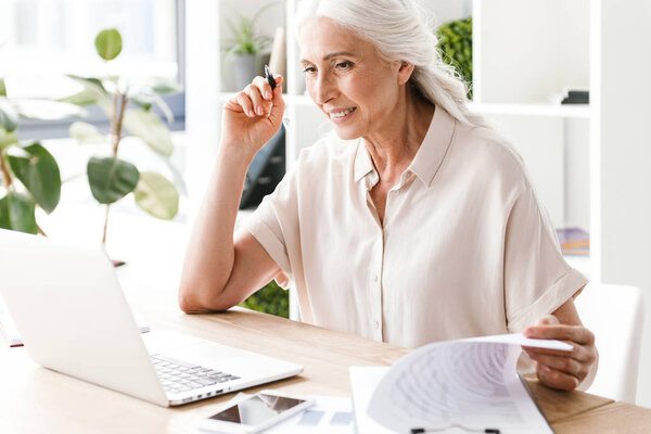Picture of mature happy cheerful woman indoors in office working writing notes.