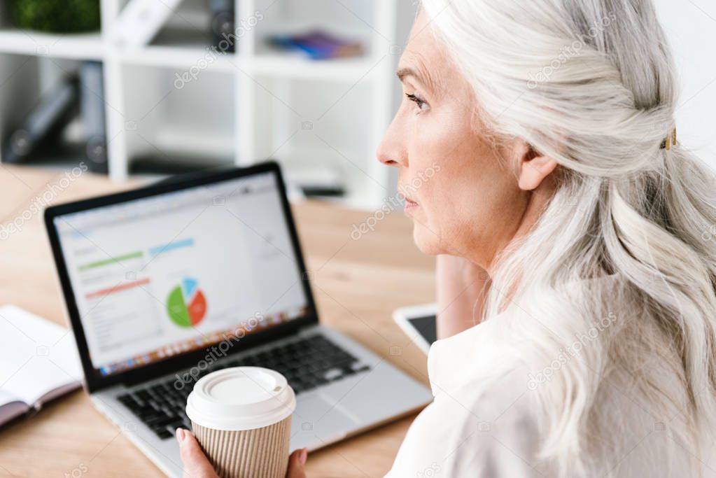 Close up of confident mature business woman working on laptop computer and drinking coffee while sitting at the desk in office