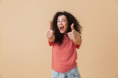 Başparmak gösterilmesini bej duvar arka plan üzerinde izole sevimli genç kadın görüntüsünü.