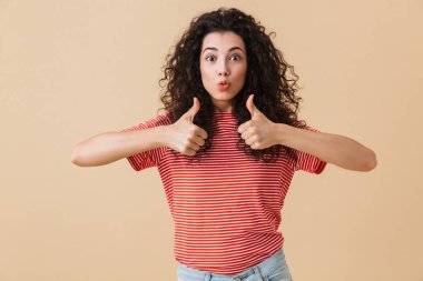 Başparmak gösterilmesini bej duvar arka plan üzerinde izole sevimli genç kadın görüntüsünü.