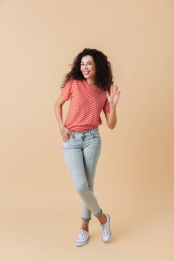 Tam boy esmer kıvırcık saçlı bej renkli arka plan üzerinde izole elini sallayarak neşeli genç bir kadın portresi