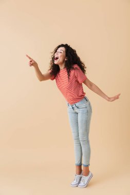 Esmer kıvırcık saçlı kopya alanı işaret heyecanlı bir genç kadın portresi tam uzunlukta bej renkli arka plan üzerinde izole