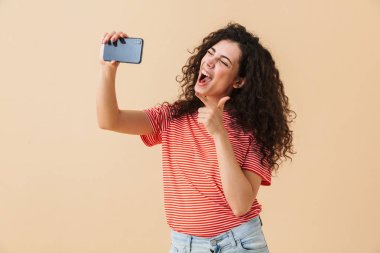 Cep telefonu işaret ederek konuşmaya selfie yapmak imge-in şaşırtıcı genç kıvırcık kadın bej renkli arka plan üzerinde izole ayakta.