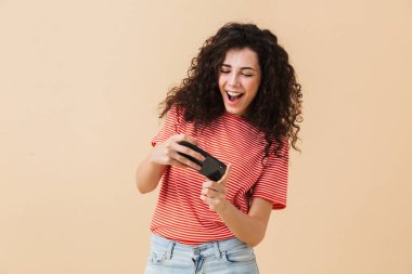 Duygusal heyecanlı genç kıvırcık kadın bej renkli arka plan üzerinde oyun oynamak cep telefonuyla izole ayakta görüntüsünü.