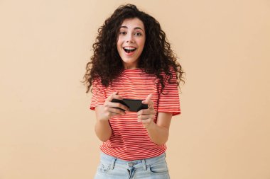 Bej renkli arka plan üzerinde oyun oynamak cep telefonuyla izole duran duygusal heyecanlı genç kıvırcık kadın görüntüsü. Görünümlü kamera.