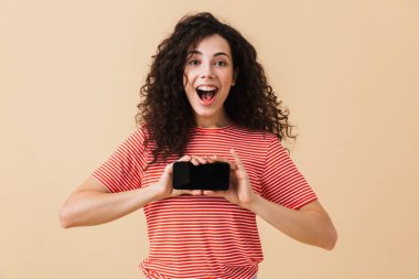 Duygusal heyecanlı genç kıvırcık kadın ayakta ekran cep telefonu tarafından gösterilen bej renkli arka plan üzerinde izole görüntüsü. Görünümlü kamera.