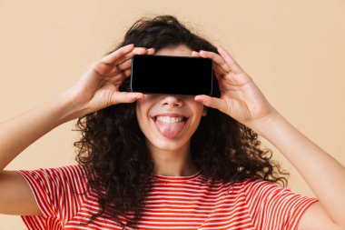 Gözleri kapsayan cep telefonuyla görüntü gösterilen bej renkli arka plan üzerinde izole duran sevimli genç kıvırcık kadın görüntüsünü.