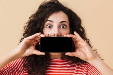Ekran cep telefonu tarafından ağız kapsayan gösterilen bej renkli arka plan üzerinde izole duran şok genç kıvırcık kadın görüntüsünü.