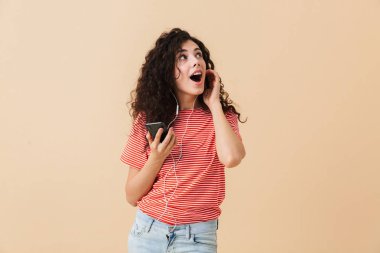 Bej renkli arka plan üzerinde izole kulaklık ile müzik dinlemek duygusal şok heyecanlı genç kıvırcık kadın görüntüsü. Bir kenara arıyorsunuz.