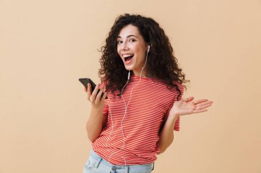 Bej renkli arka plan şarkı üzerinde izole kulaklık ile müzik dinlemek duygusal mutlu heyecanlı genç kıvırcık kadın görüntüsünü.