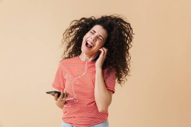 Bej renkli arka plan üzerinde dans izole kulaklık ile müzik dinlemek duygusal mutlu heyecanlı genç kıvırcık kadın görüntüsünü.