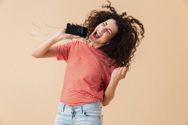 Bej renkli arka plan üzerinde izole kulaklık ile müzik dinlemek duygusal mutlu heyecanlı genç kıvırcık kadın görüntüsünü.