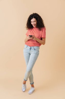 Cep telefonuyla bej renkli arka plan üzerinde izole gülümseyen bir casual kız portresi tam uzunlukta
