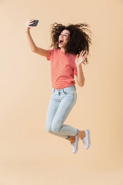 Tam uzunlukta atlama ve sallayarak bej renkli arka plan üzerinde izole ederken cep telefonu ile bir selfie alarak neşeli bir genç kız portresi