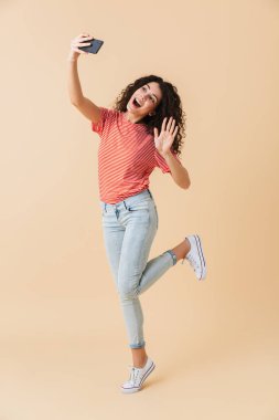 Tam uzunlukta atlama ve sallayarak bej renkli arka plan üzerinde izole ederken cep telefonu ile bir selfie alarak neşeli bir genç kız portresi