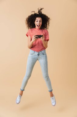 Tam uzunlukta portre neşeli bir genç kızın atlama bej renkli arka plan üzerinde izole süre iskambil spor üstünde hareket eden telefon