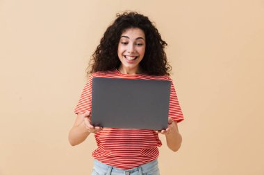 Dizüstü bilgisayar kullanan bej renkli arka plan üzerinde izole şaşırtıcı oldukça genç kıvırcık kadın görüntüsünü.