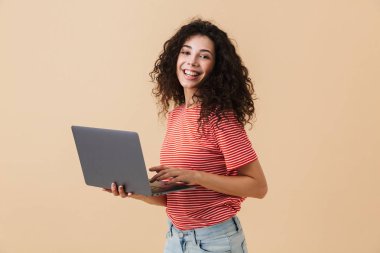 Dizüstü bilgisayar kullanan bej renkli arka plan üzerinde izole heyecanlı genç ve güzel kıvırcık kadın görüntüsü. Görünümlü kamera.