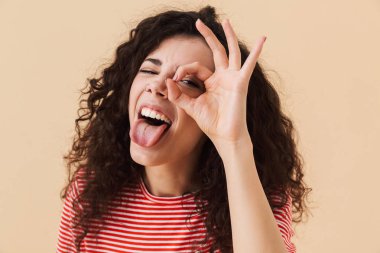 İyi jest yapmak imge-in duygusal genç kıvırcık kadın kamera seyir bej renkli arka plan üzerinde izole.