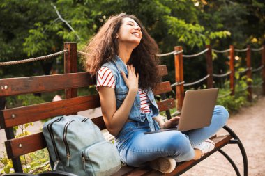 Yaz gününde yeşil parkta bankta oturmuş ve istimal gümüş laptop sırt çantası ile memnun rahat kadın görüntüsünü