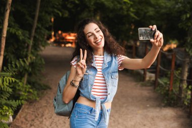 Esmer güzel kadın 18-20. barış işareti ve alma selfie Smartphone cep telefonu ile duran yeşil park yolundaki gösterilen sırt çantası ile fotoğrafı