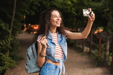 Esmer güzel kadın 18-20 geniş gülümsüyor ve yeşil park yol boyunca yürürken cep telefonundan selfie fotoğrafı çekmek sırt çantası ile fotoğrafı