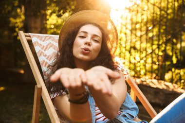 Güzel baştan çıkarıcı kadın 18-20 hava üfleme hasır şapka için güneşli Park dinlenme sırasında lounge sandalyede otururken öp