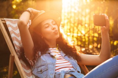 Açık havada park yapmak selfie öpücük üfleme cep telefonuyla konuşan genç güzel bir kıvırcık kadın görüntüsünü.