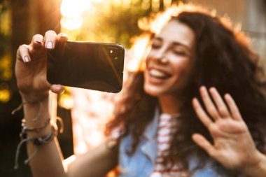 Güneşli günde 18-20 gülümseyen ve sırasında selfie fotoğraf çekerken cep telefonu arıyorum şezlong Park içinde dinlenme sevimli kıvırcık kadın portresi