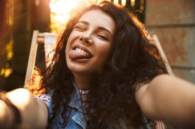 Dil gösterilen kamera tarafından selfie yapmak imge-in komik genç güzel kıvırcık kadın açık havada park.