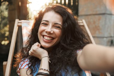 Park lounge sandalyede oturan güneşli günde 18-20 kameraya bakarak ve selfie alarak mutlu neşeli öğrenci kız fotoğraf