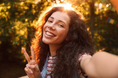 Sevimli kıvırcık kız 18-20 arıyorsunuz kameraya gülümseyen ve zafer gösterilen açık selfie fotoğraf çekerken oturum açın